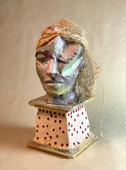 A unique mixed media sculpture made from stoneware clay, with vibrant paint and twine accents, depicting a human face.