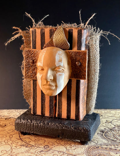 A mixed media sculpture with a ceramic face mask, burlap, and striped materials, mounted on a wooden base.