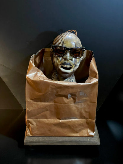 Metallic ceramic sculpture with sunglasses inside a brown paper bag on a dark background