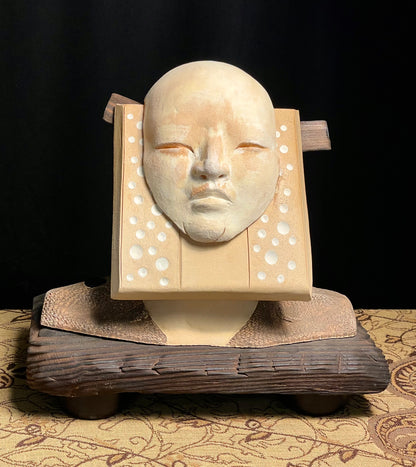 A sculptural sundial featuring a stylized face with a wooden frame and stoneware clay details, mounted on a wooden base with a patterned fabric backdrop.