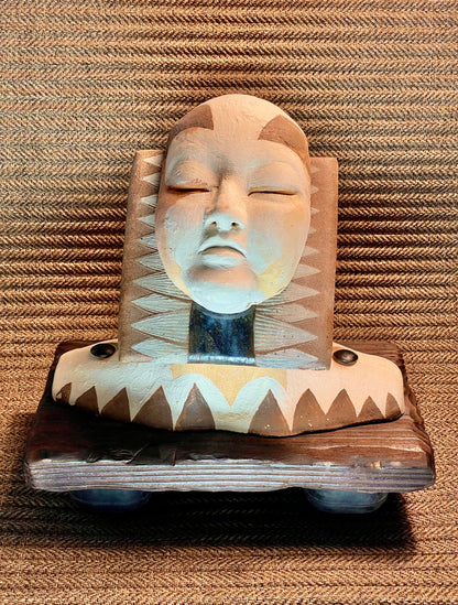 A sculptural artwork depicting a stylized face with closed eyes, featuring geometric patterns and a mix of materials, placed on a woven surface.