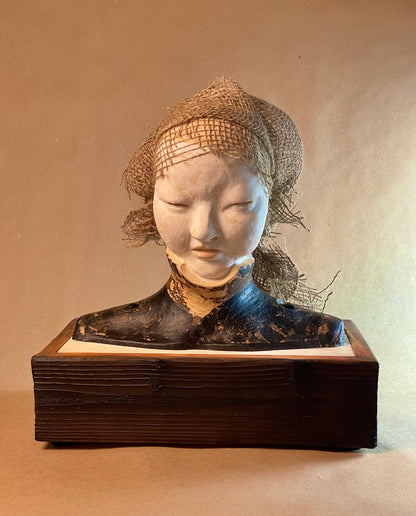 A sculptural artwork of a female figure with a textured hat, made from materials including stoneware clay, wood, burlap, paint, and metal, displayed on a wooden surface against a beige background.