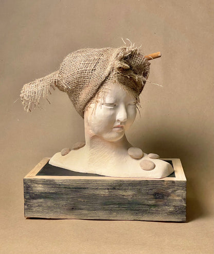 A sculpture featuring a head with a burlap covering, set against a wooden base with a neutral background.