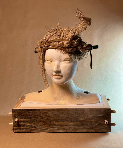 A sculpture featuring a human-like face with a textured, decorative headpiece made from materials such as stoneware clay, wood, and burlap.