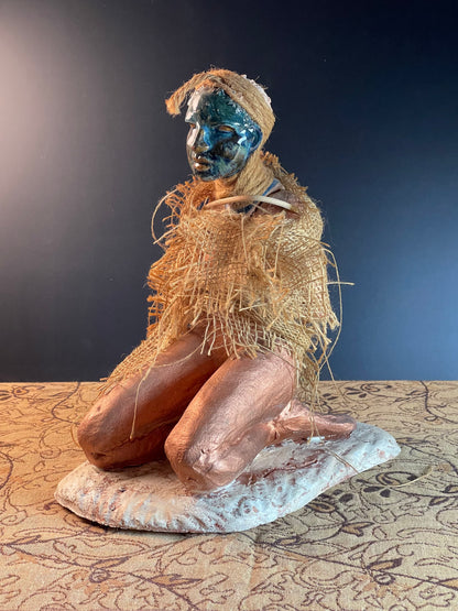 A sculpture of a seated figure with a naturalistic style, featuring a combination of stoneware clay, glaze, paint, bamboo, and burlap materials. The figure appears to be wearing a grassy, rustic garment and is set against a patterned background.