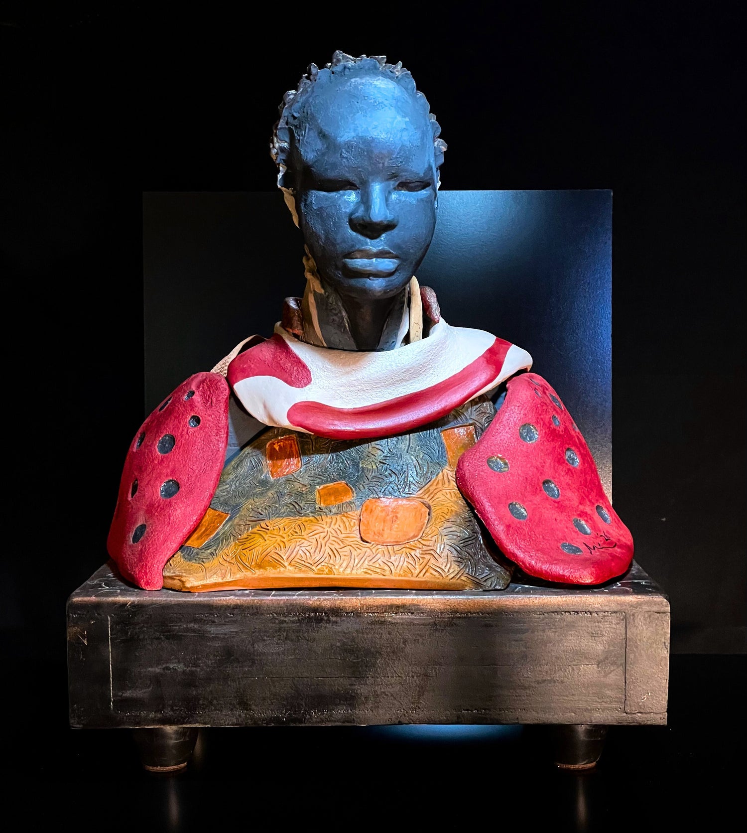 Sculpture of a bust with blue face, red and white garment, and textured surface on a dark background